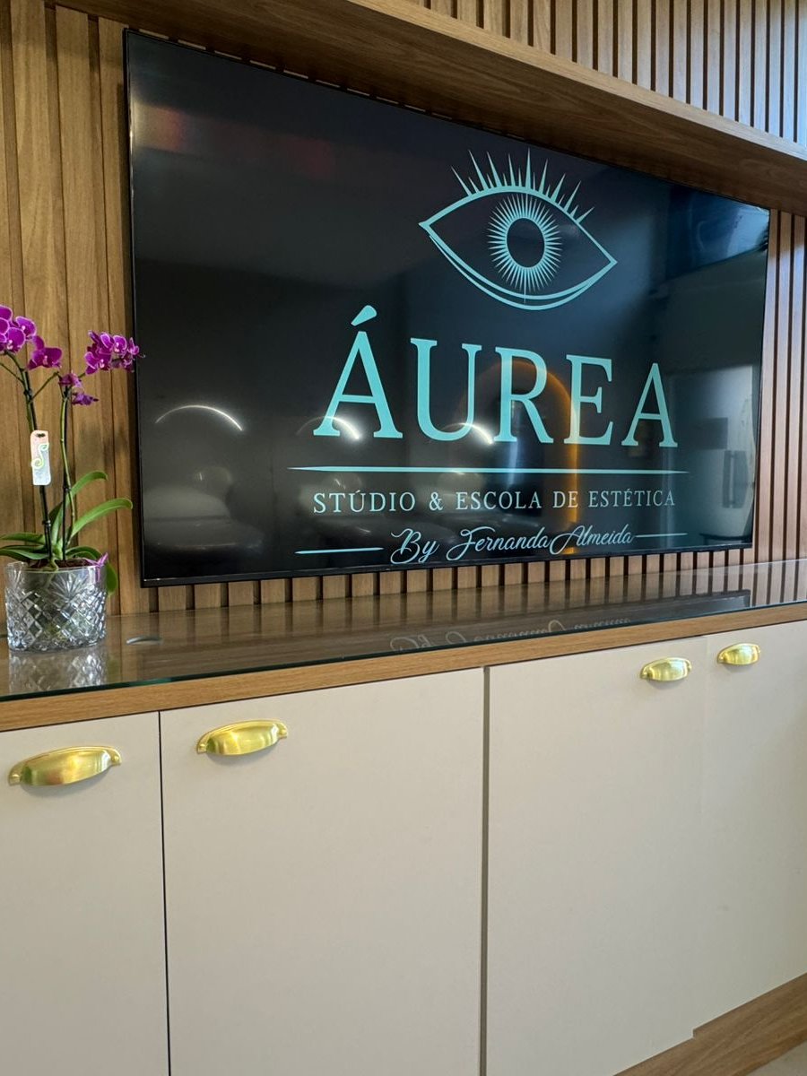 Sala de formação da Áurea, em Resende, com painel ripado e TV exibindo a marca, onde acontece o curso de Alongamento de Cílios