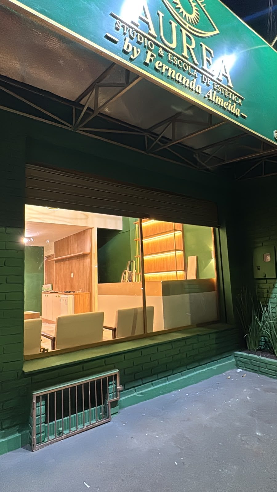 Fachada da Áurea à noite, com letreiro aceso e recepção iluminada vista pela vitrine